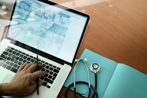 Hand typing on laptop keyboard next to a stethoscope 